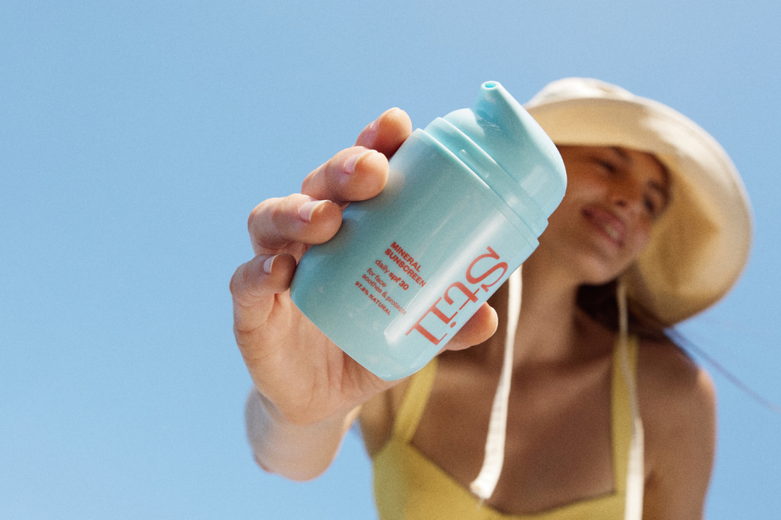 Bottle of Stil Suncare in the foreground of the image, behind a woman smiling with a hat on