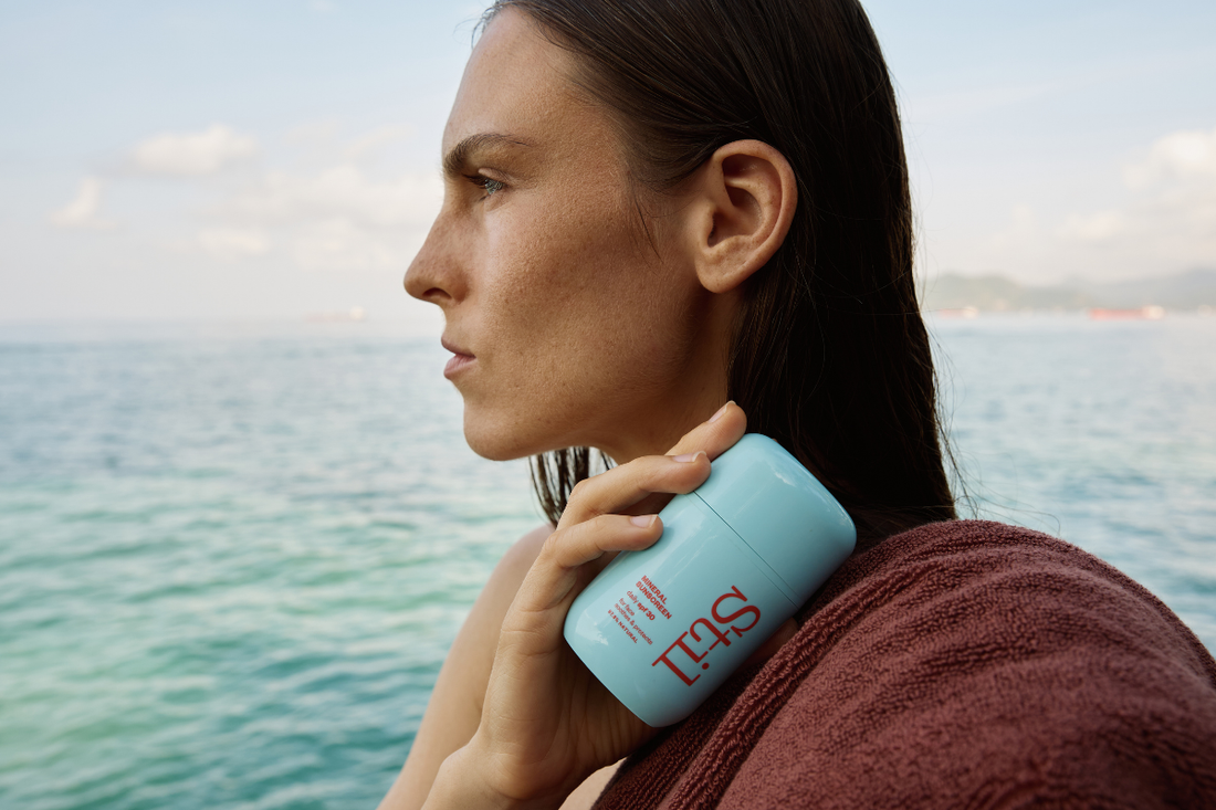 Woman holding a bottle of Stil looking out at the sea with a towel over her shoulder