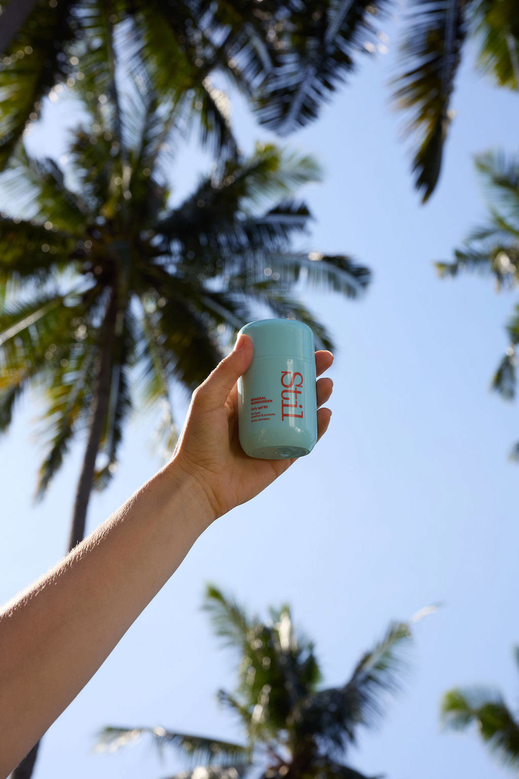 A hand holding a small blue Stil Suncare bottle with a bikini red logo, set against a background of palm trees and a clear blue sky.