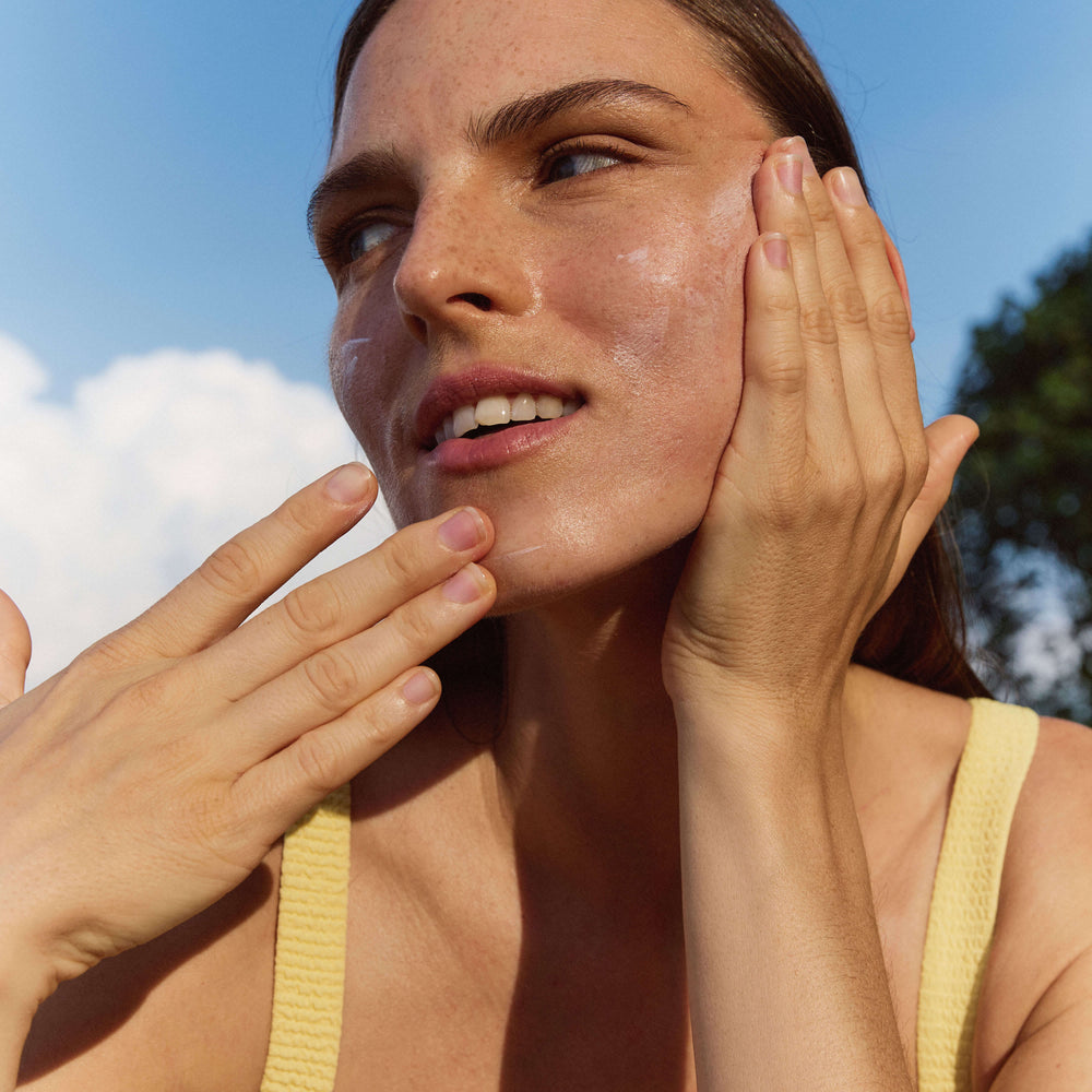 A close-up outdoor shot of a woman with her hands touching her face, wearing a pale yellow ribbed tank top against a blue sky background.