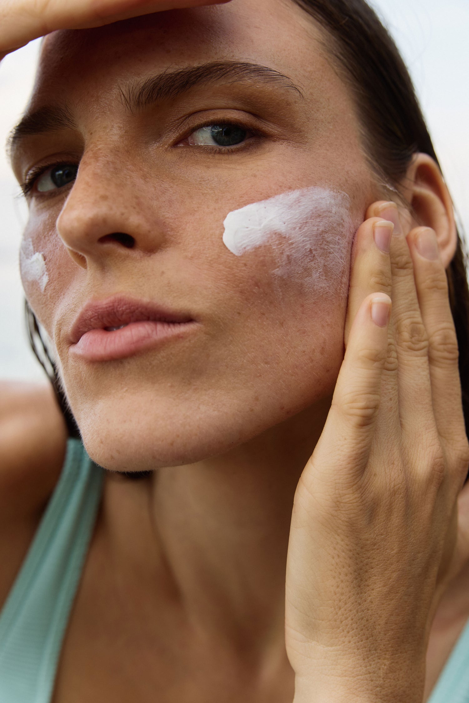 Close-up of a woman applying mineral sunscreen cream to her face, wearing a light teal tank top against a bright outdoor background.