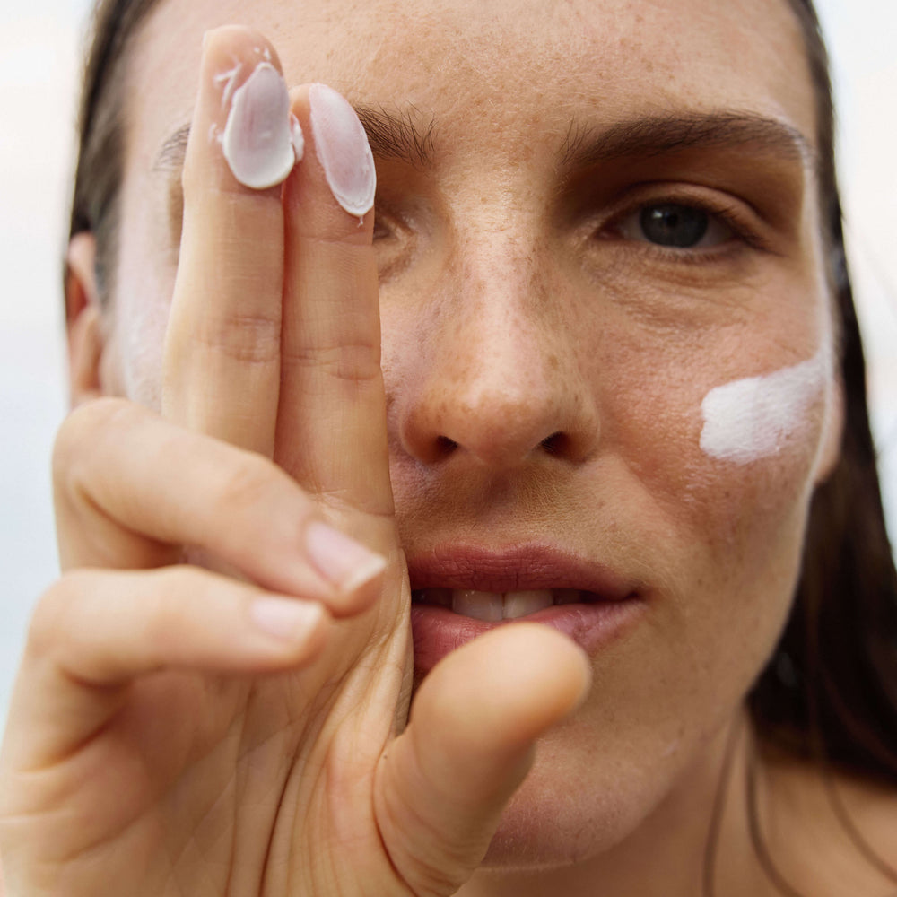 Close-up of a woman applying or showing white sunscreen cream on her cheek and holding her fingers near her face, with cream visible on her fingertips.